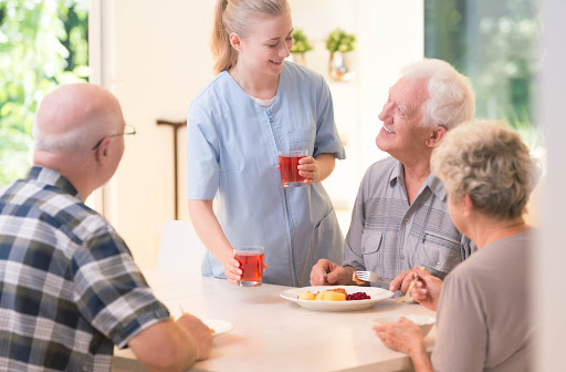 A nurse holds two glasses of juice while standing over a table of three smiling seniors eating. The nurse is handing juice to one of the seniors.