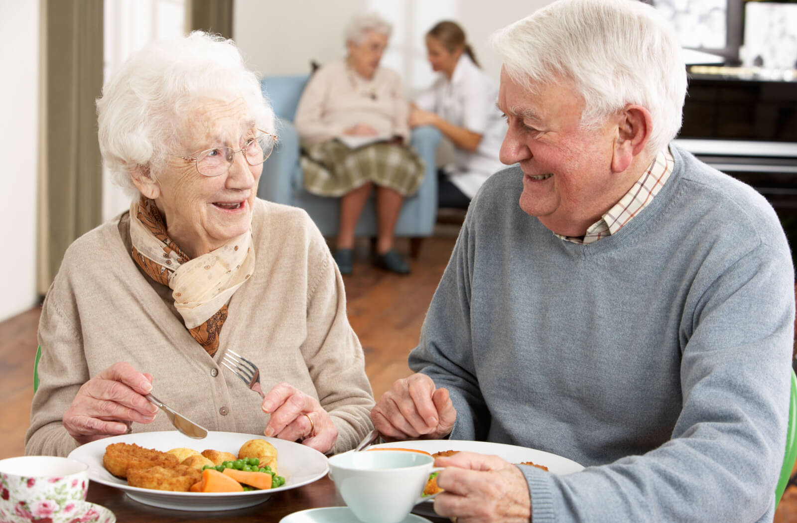 A senior woman and senior man smiling and sitting at a table together eating breakfast and drinking tea.