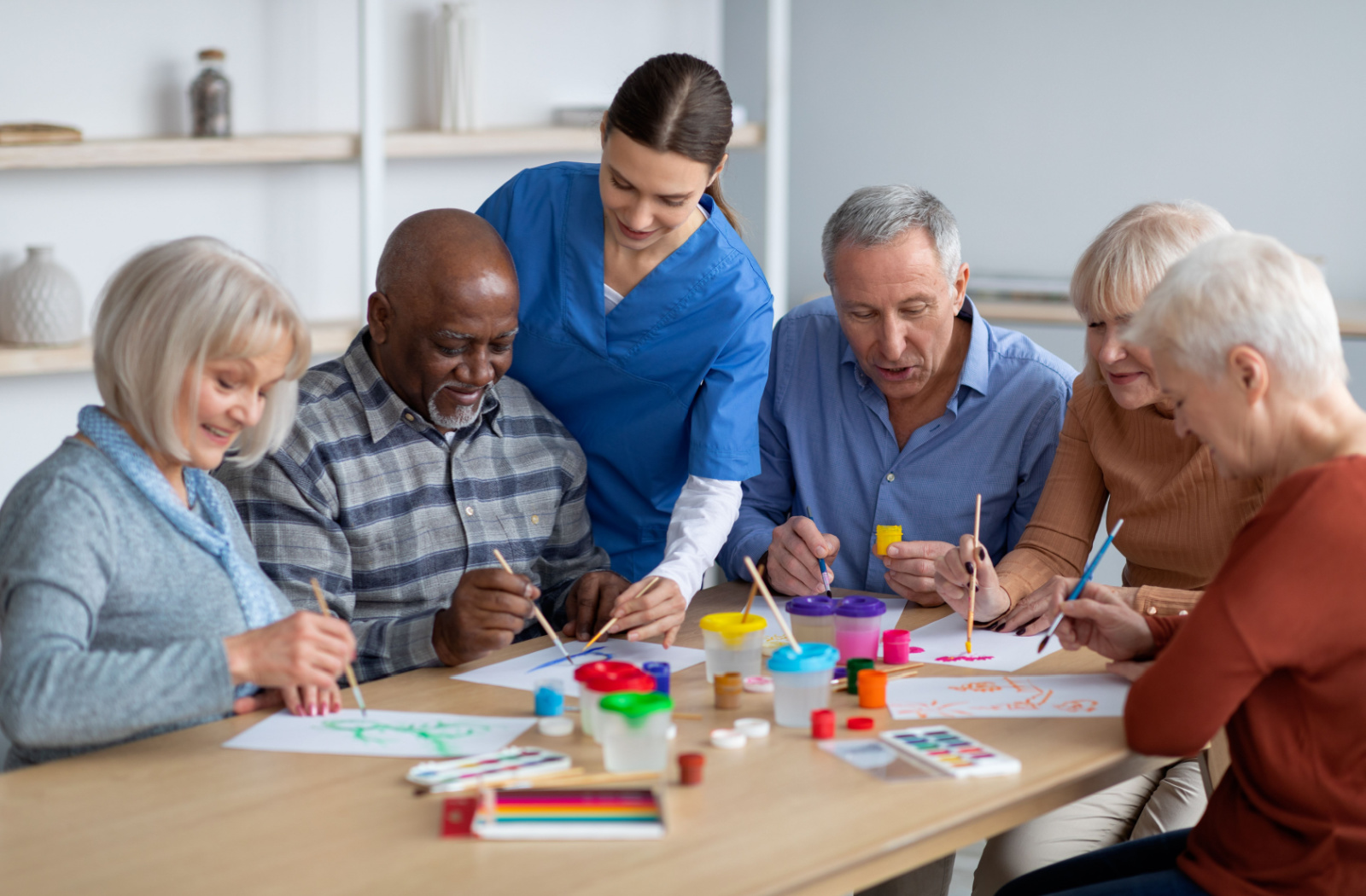 A group of seniors sitting at a table holding small flat brushes and painting, while being supervised by a nurse.