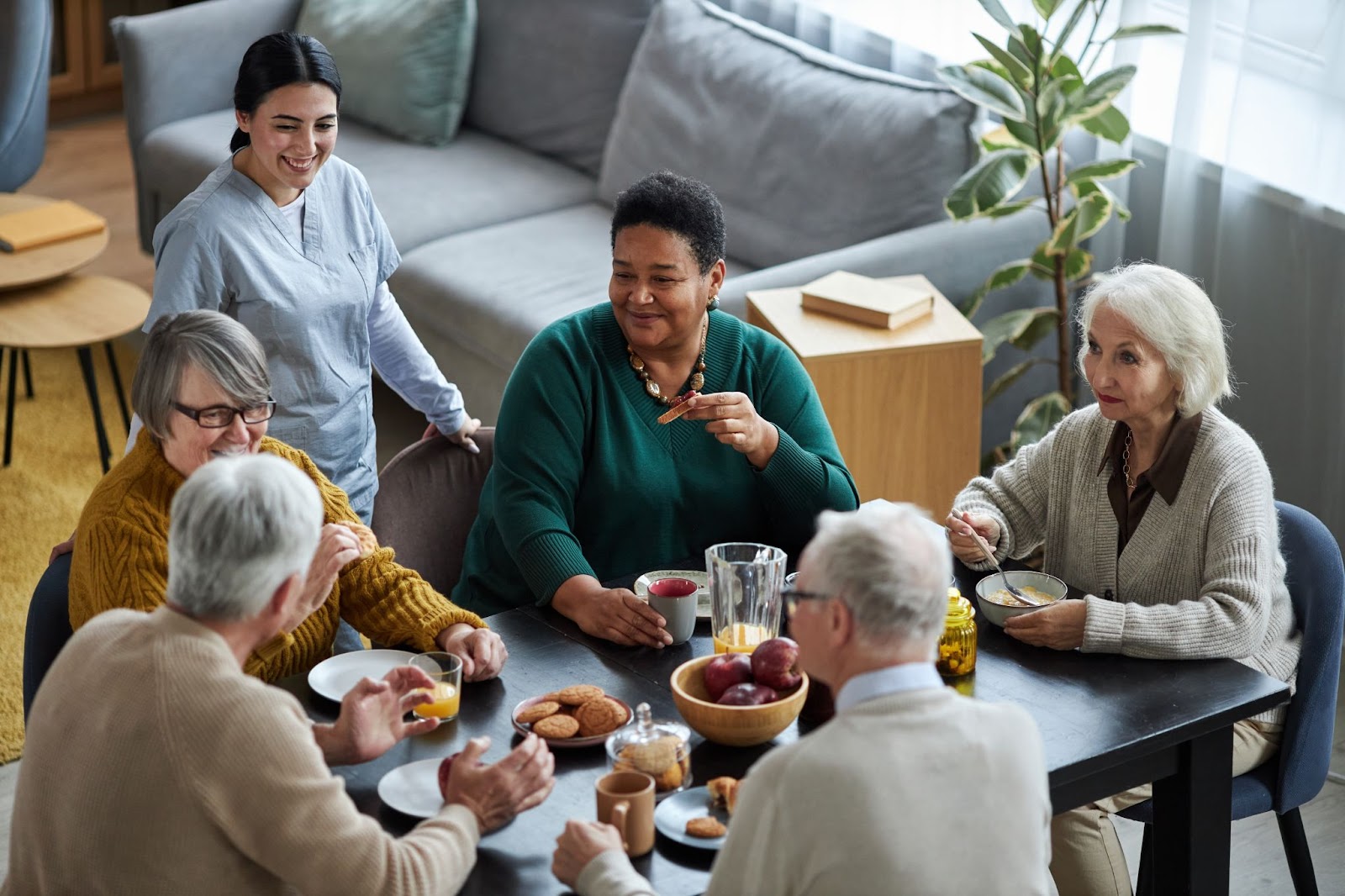 A group of seniors and their caretaker sit around a table sharing a meal together.