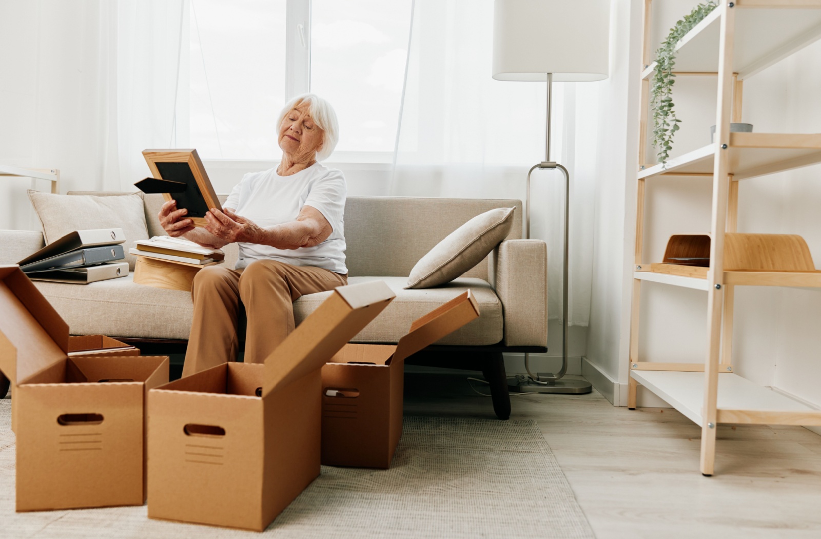 A woman on moving day surrounded by cardboard boxes, holding and looking at a framed photo with a thoughtful expression.