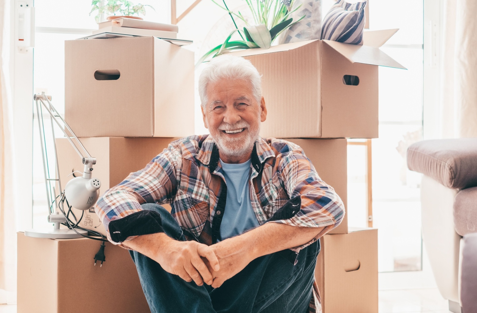A smiling older adult sitting on the ground in front of stacked moving boxes preparing to move to assisted living.