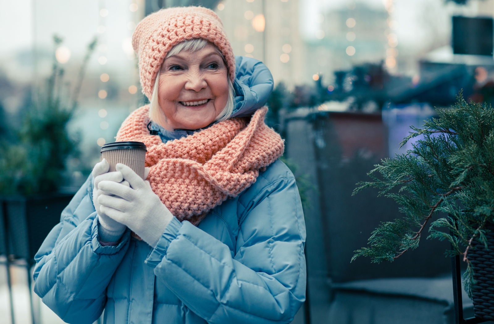 An older woman dressed warmly in a knit hat and scarf, smiling and holding a cup of coffee outdoors during the winter.
